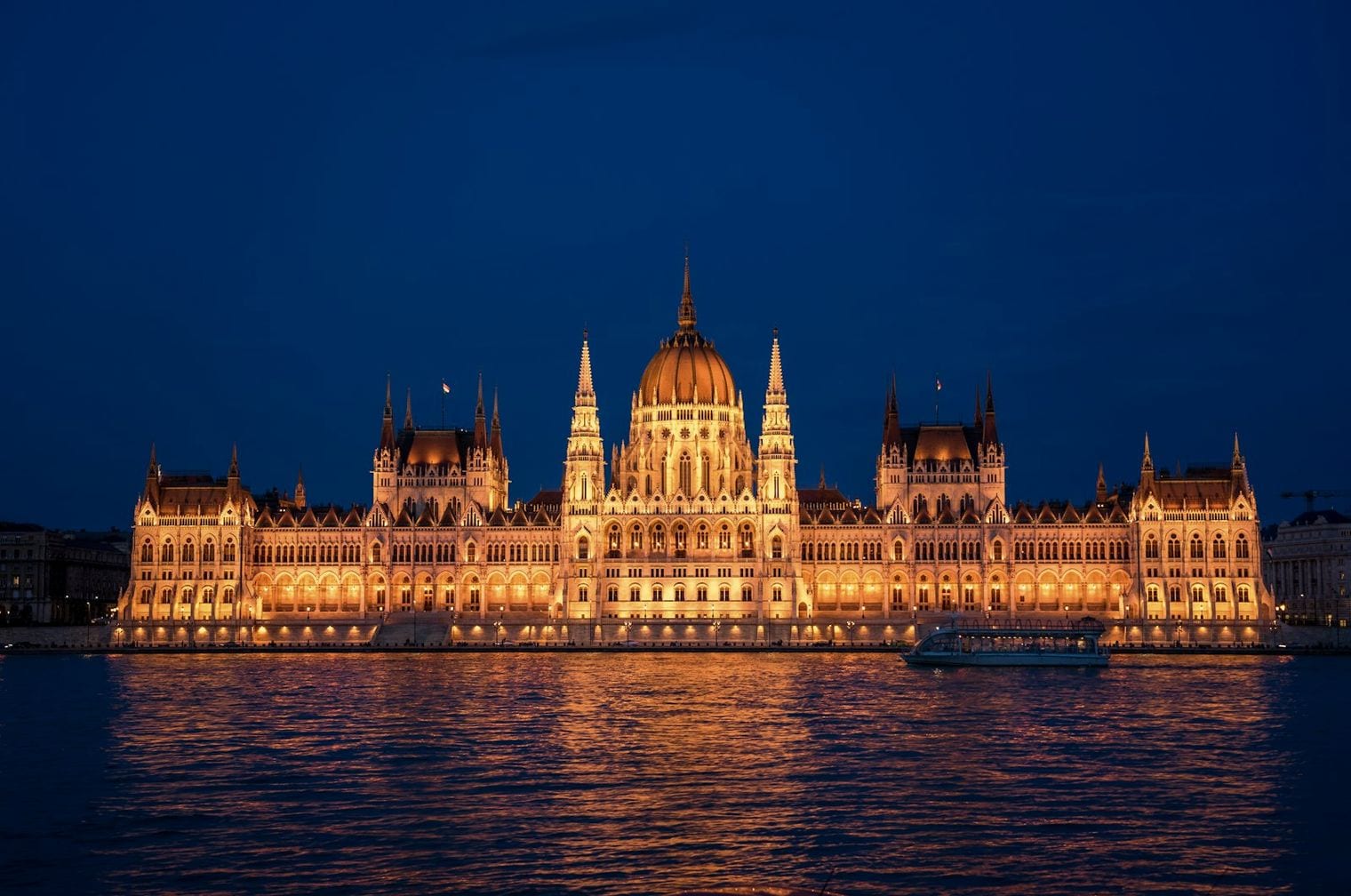The Hungarian Parliament Building lit gold at night on the bank of the Danube in Budapest, with a river cruise boat passing on the dark water.
