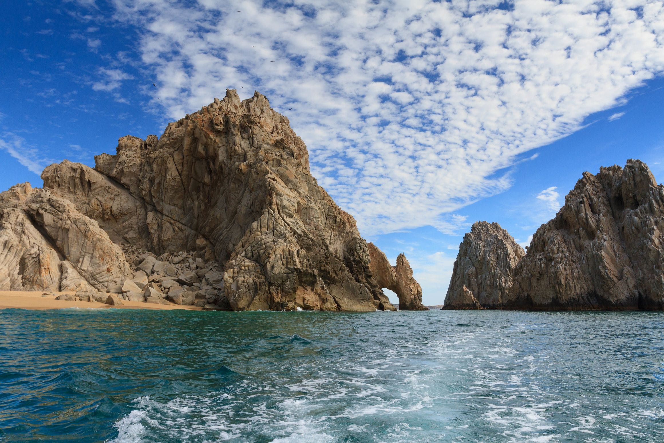 The granite sea arch at Land's End, seen from the water with the pale sand of Lovers Beach beside it and turquoise sea in the foreground.