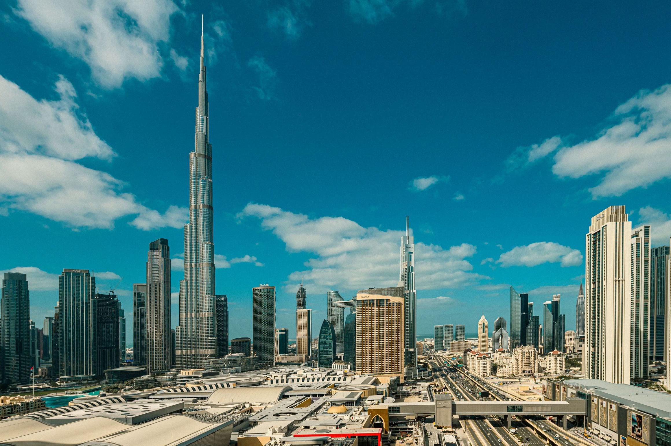The Burj Khalifa rising above the Downtown Dubai skyline on a clear day, with the roofs of the Dubai Mall below and white cloud over the city.