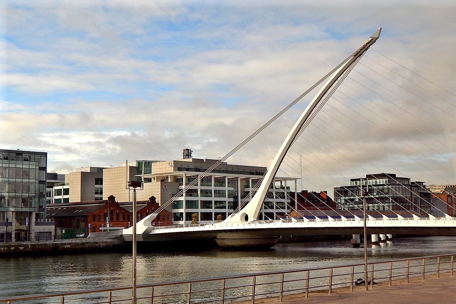 The harp-shaped white pylon and cables of the Samuel Beckett Bridge spanning the River Liffey, with the Dublin docklands quays behind