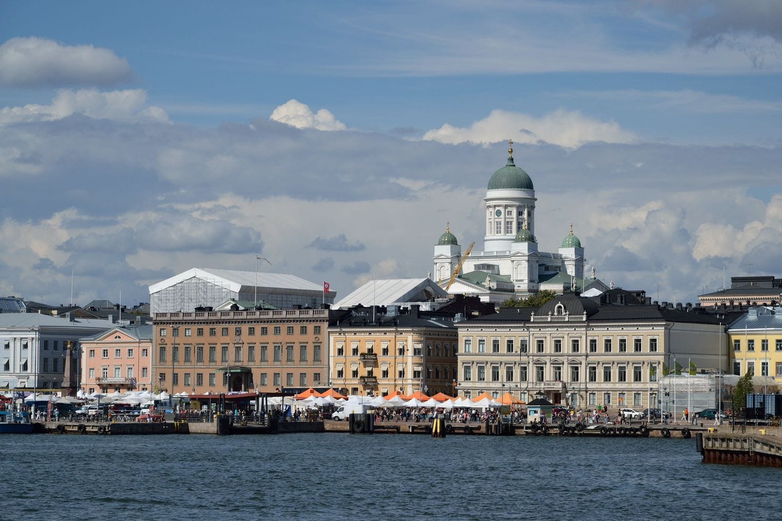 Market stalls under white and orange awnings line the Helsinki harbour front, with pastel neoclassical facades and the green dome of Helsinki Cathedral rising behind.
