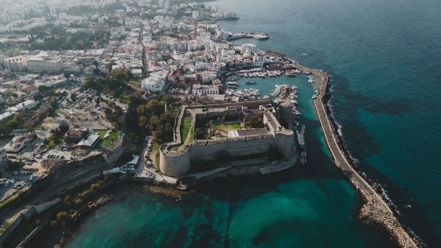 Aerial view of the horseshoe harbour at Kyrenia in northern Cyprus, with boats moored inside the breakwater and the stone castle on the headland beside it.