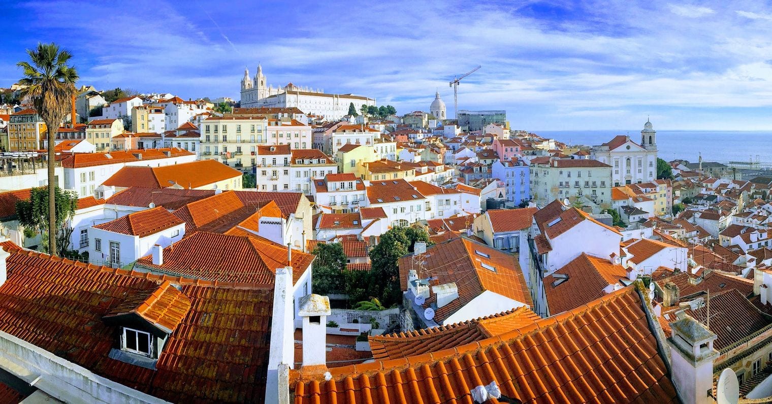 Terracotta rooftops of the Alfama district falling away toward the Tagus, with the white towers of Sao Vicente de Fora and the dome of the National Pantheon on the skyline