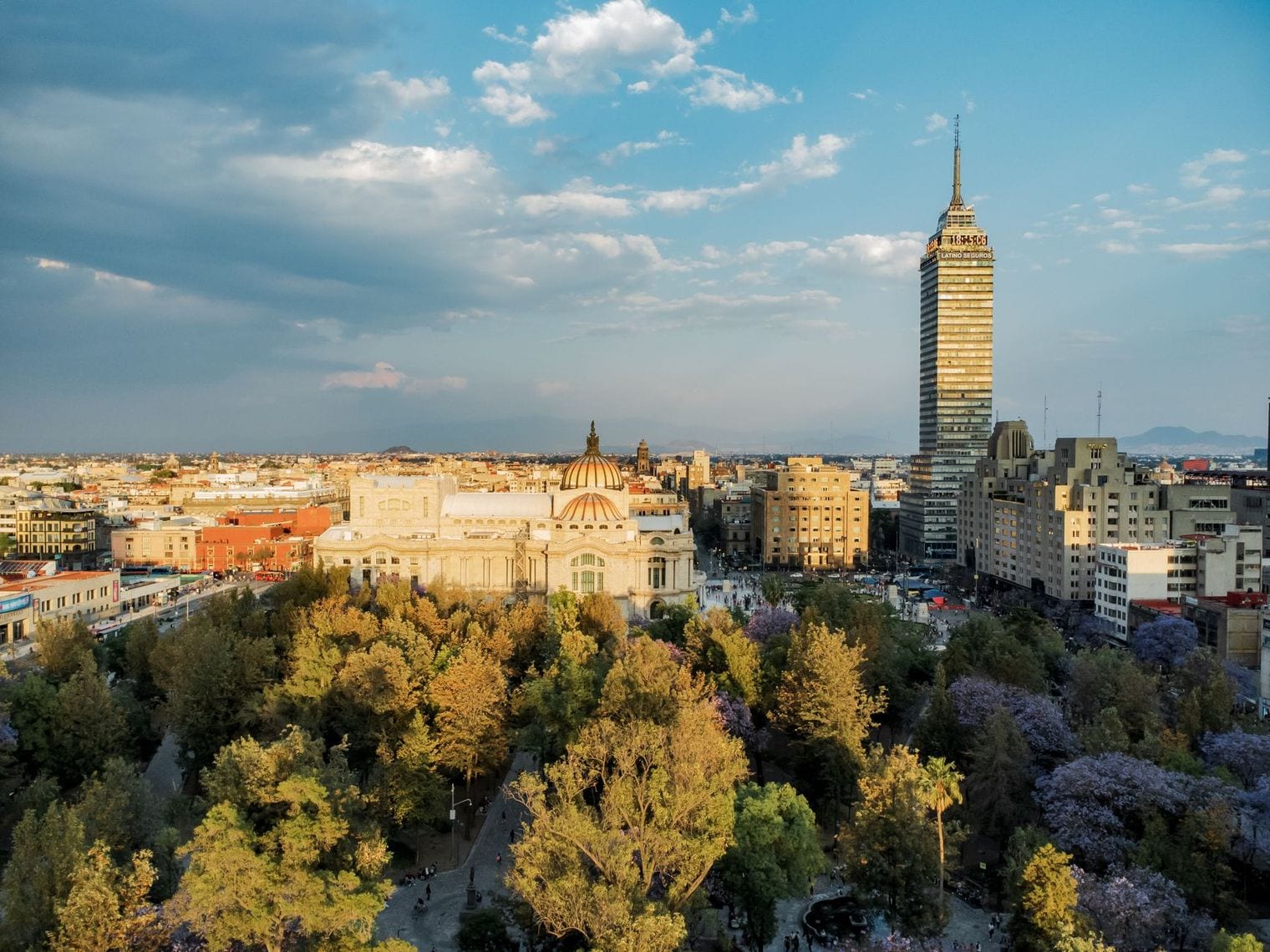 Aerial view over the trees of Alameda Central at golden hour, with the tiled dome of the Palacio de Bellas Artes below and the Torre Latinoamericana rising behind it