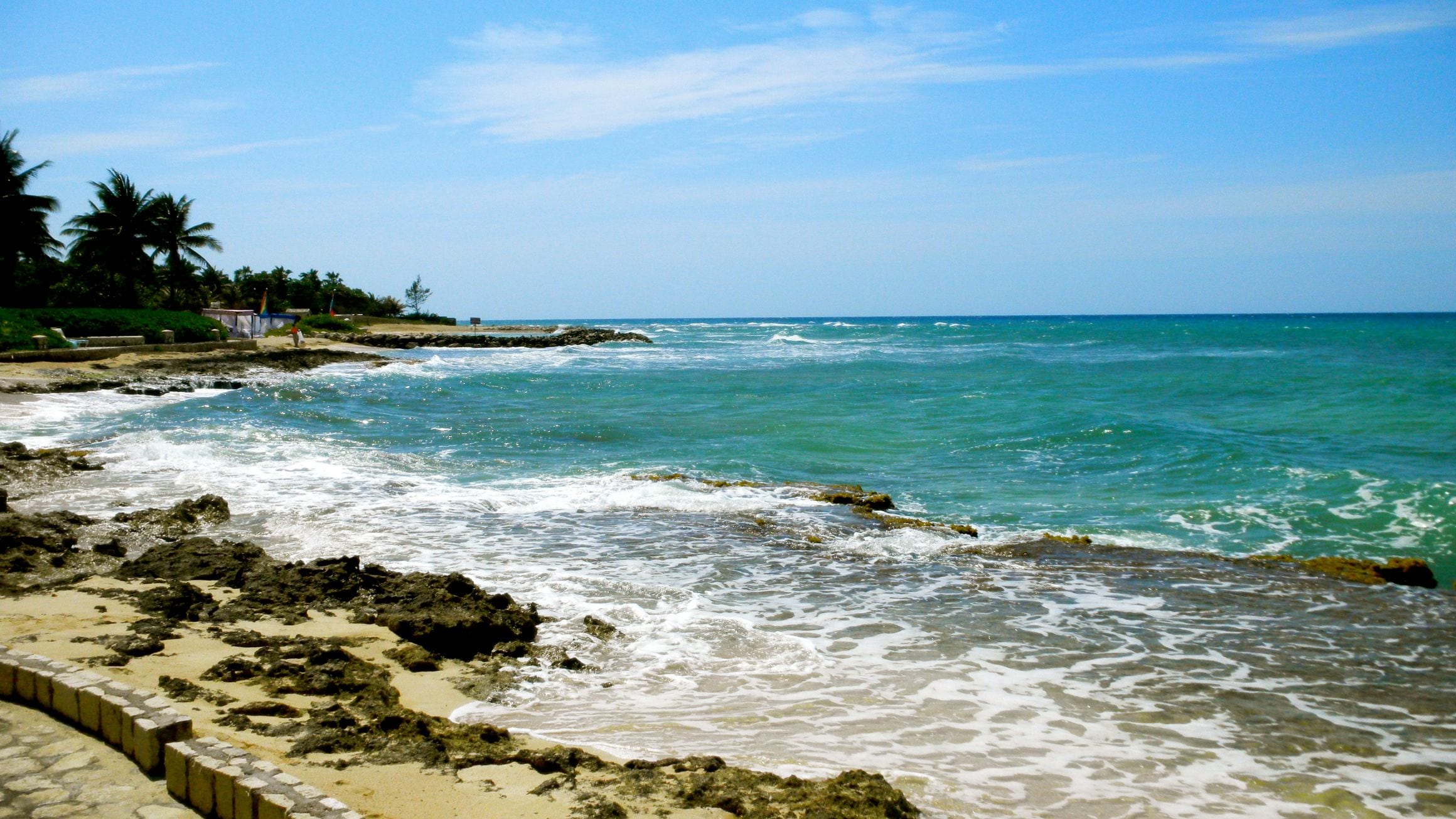 Turquoise Caribbean water breaking over a low rocky shoreline on the Montego Bay coast, with palms and a small building among greenery on the headland behind