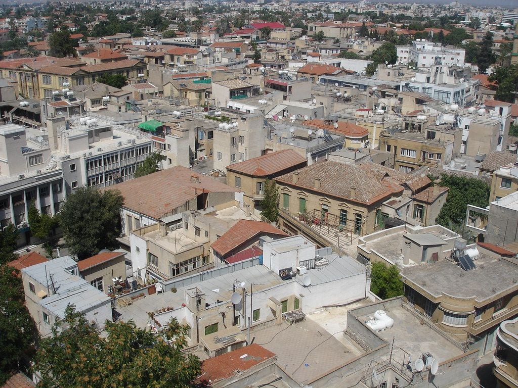 A high view across the densely packed rooftops of central Nicosia, red clay tiles and flat white roofs with water tanks running to low hills on the horizon