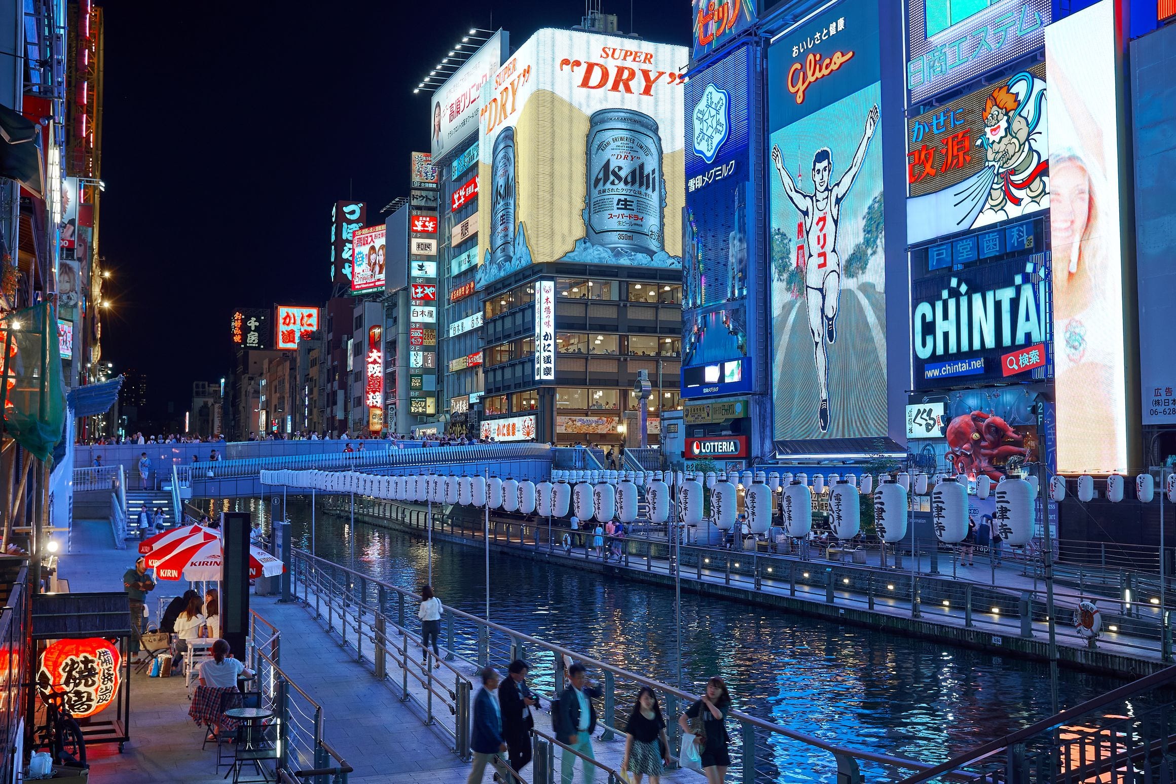 Neon signs and illuminated billboards reflected in the Dotonbori canal in Osaka after dark, with crowds along the waterside walkway.
