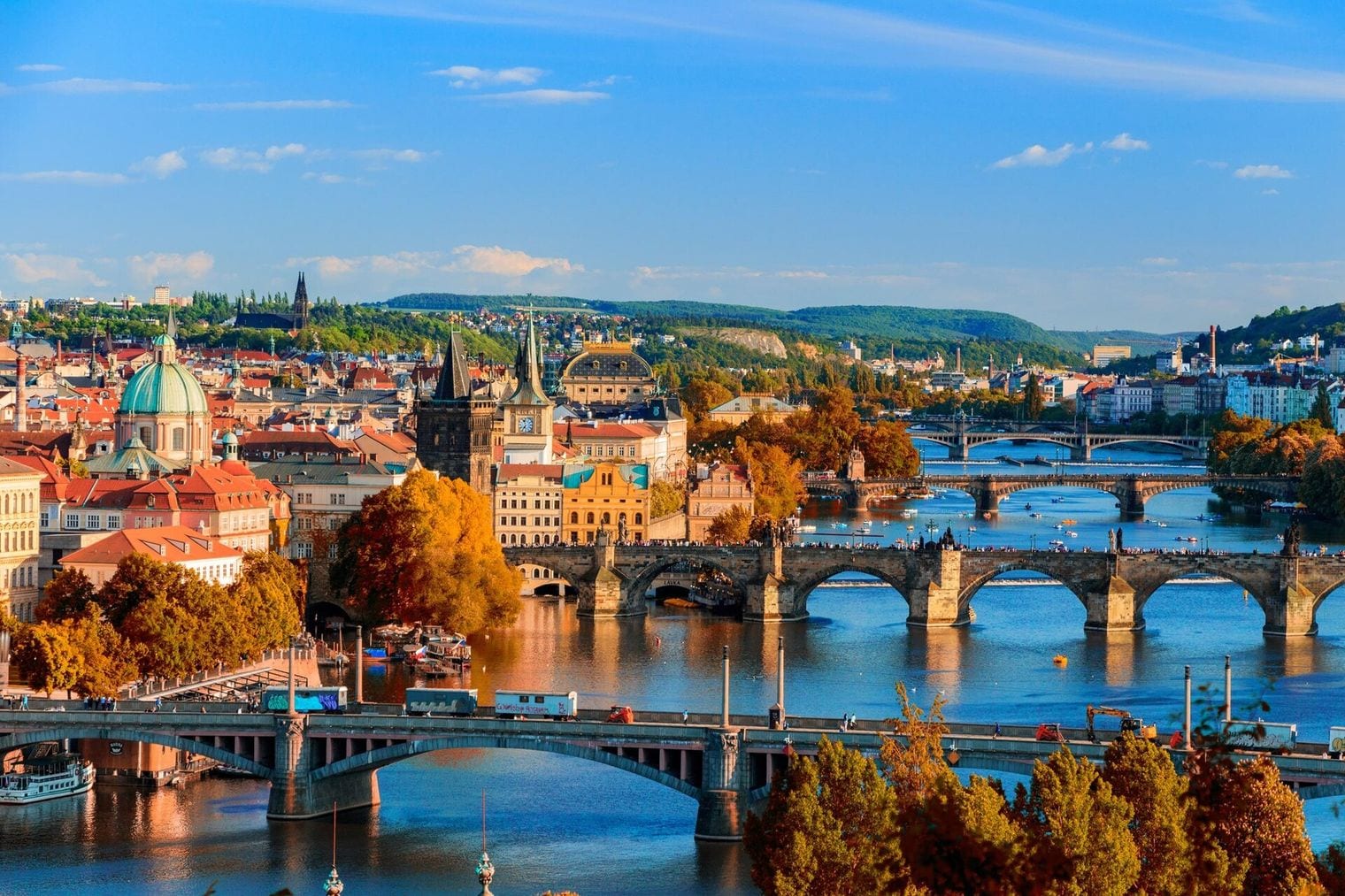 Autumn view across the Vltava in Prague with Charles Bridge and its stone arches at the centre, a chain of further bridges upstream, the red-tiled roofs and green church dome of the Old Town behind, and golden trees along both embankments