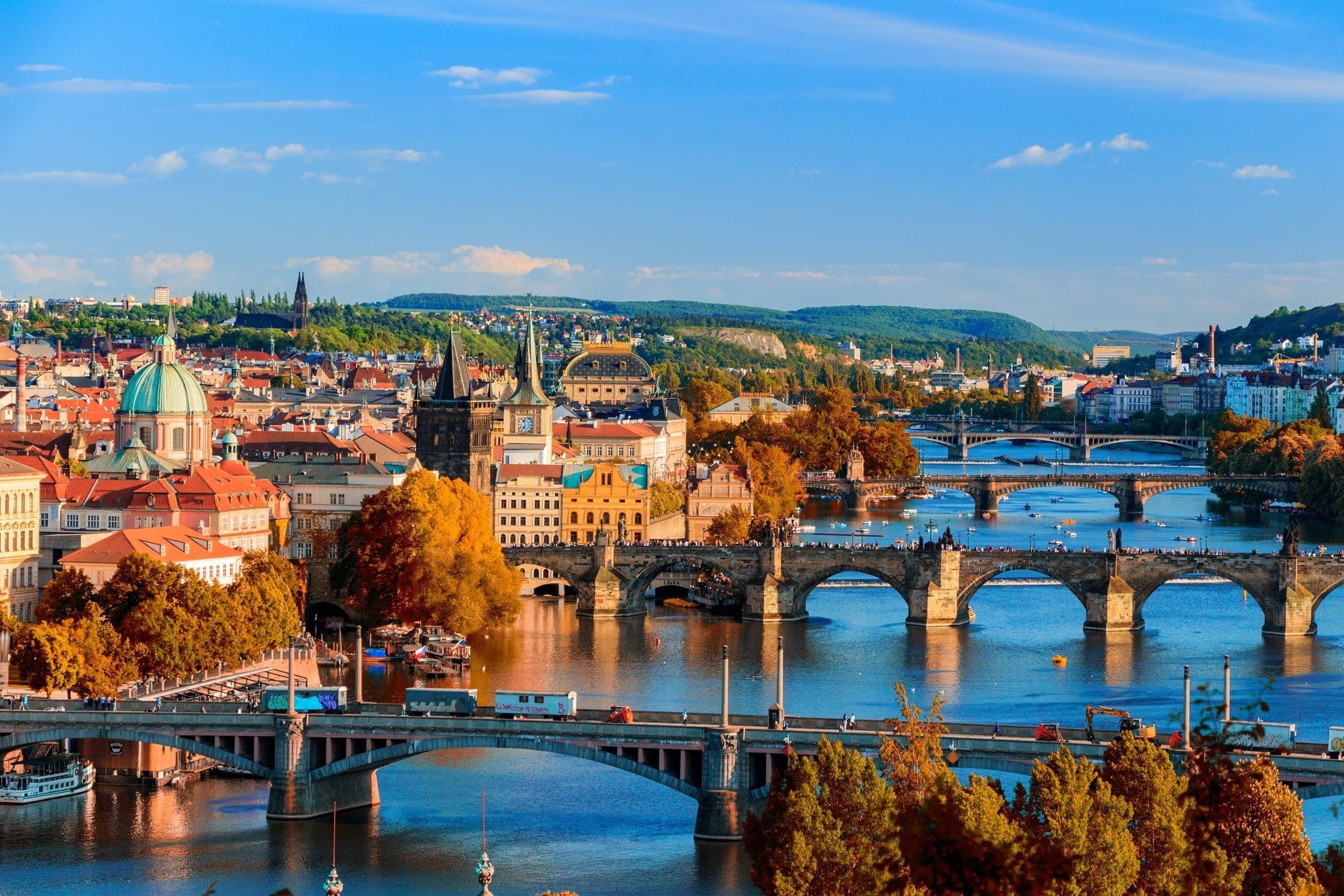 Autumn view across the Vltava in Prague with Charles Bridge and its stone arches at the centre, a chain of further bridges upstream, the red-tiled roofs and green church dome of the Old Town behind, and golden trees along both embankments