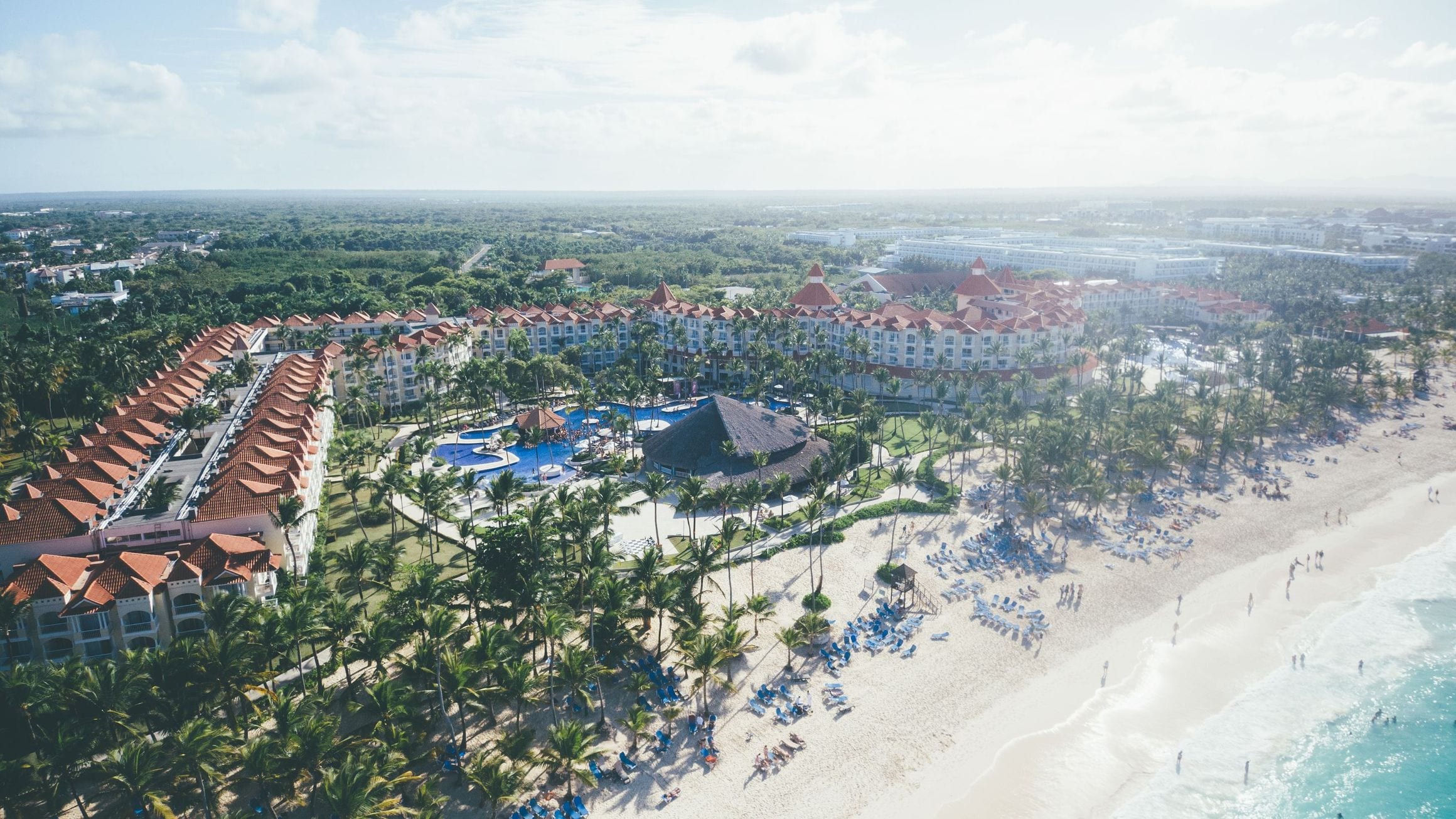 Aerial view over the Punta Cana resort strip, with low-rise hotels and palm gardens behind a curve of white sand and turquoise Caribbean water.