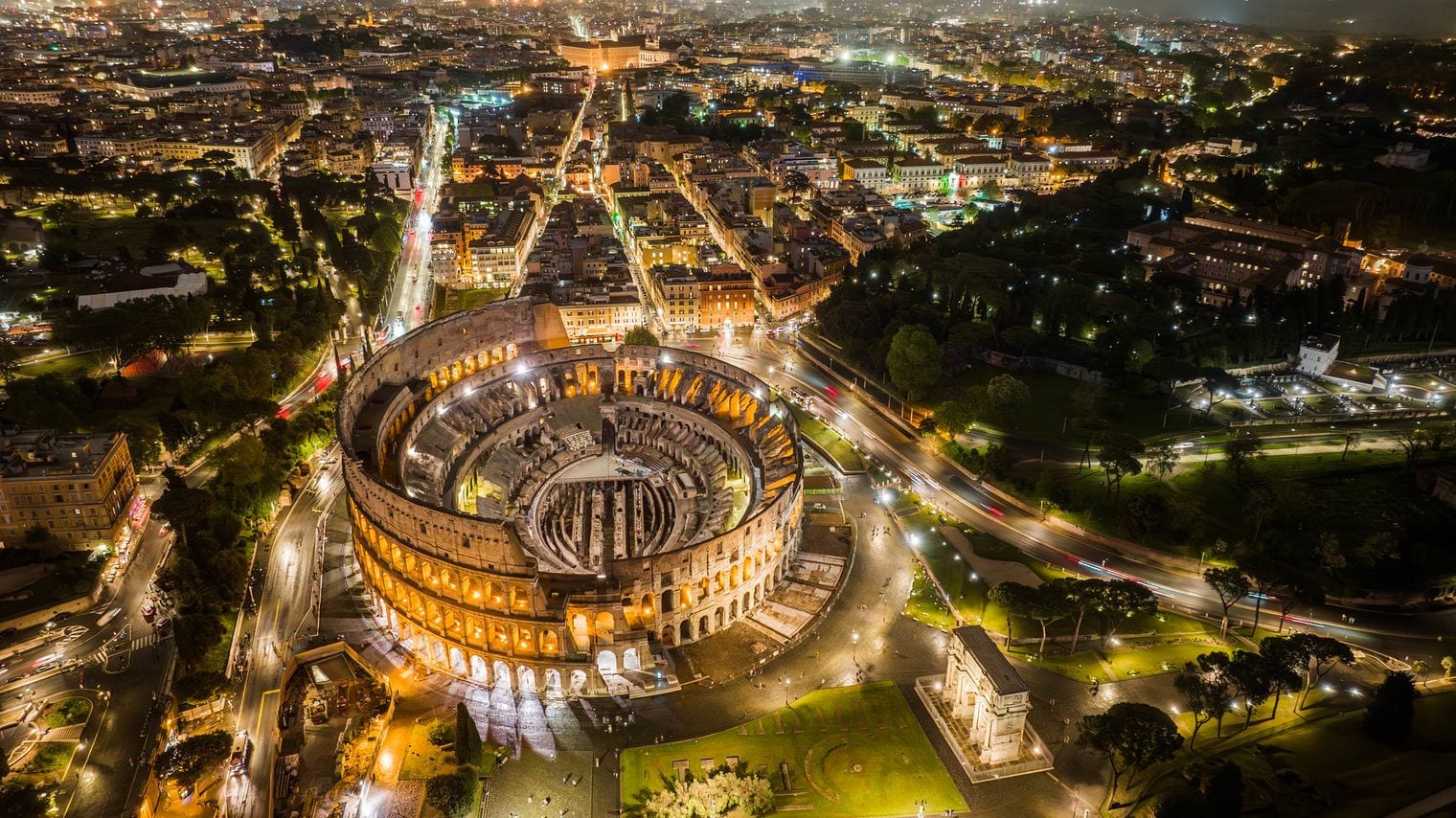 Aerial view of the illuminated Colosseum at night, with the Arch of Constantine beside it and the lit streets of central Rome spreading out behind