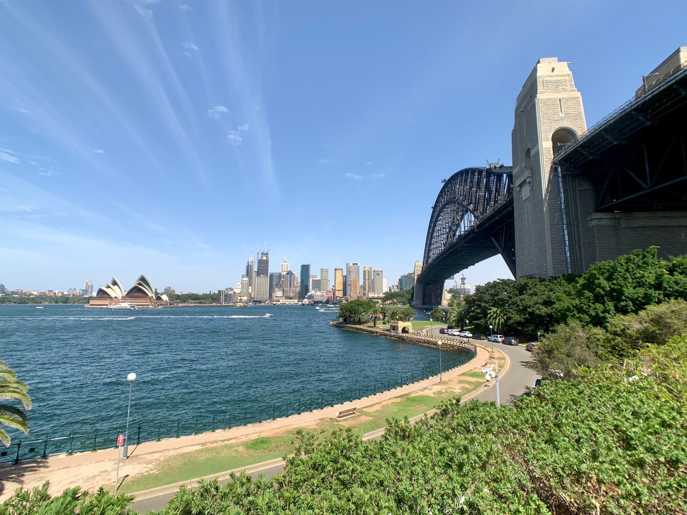 Sydney Harbour seen from the north shore, with the white shells of the Opera House and the city towers across the water and the steel arch and granite pylon of the Harbour Bridge rising at the right.