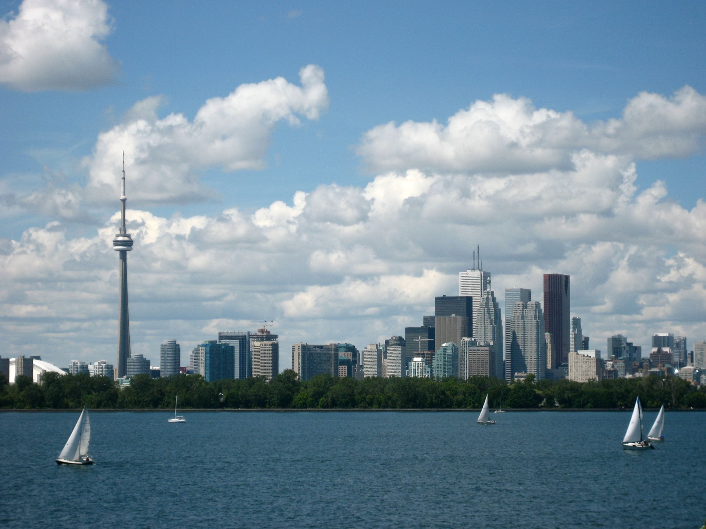 Sailboats on Lake Ontario with the Toronto skyline behind them, the CN Tower rising above the downtown office towers under a cloudy summer sky.