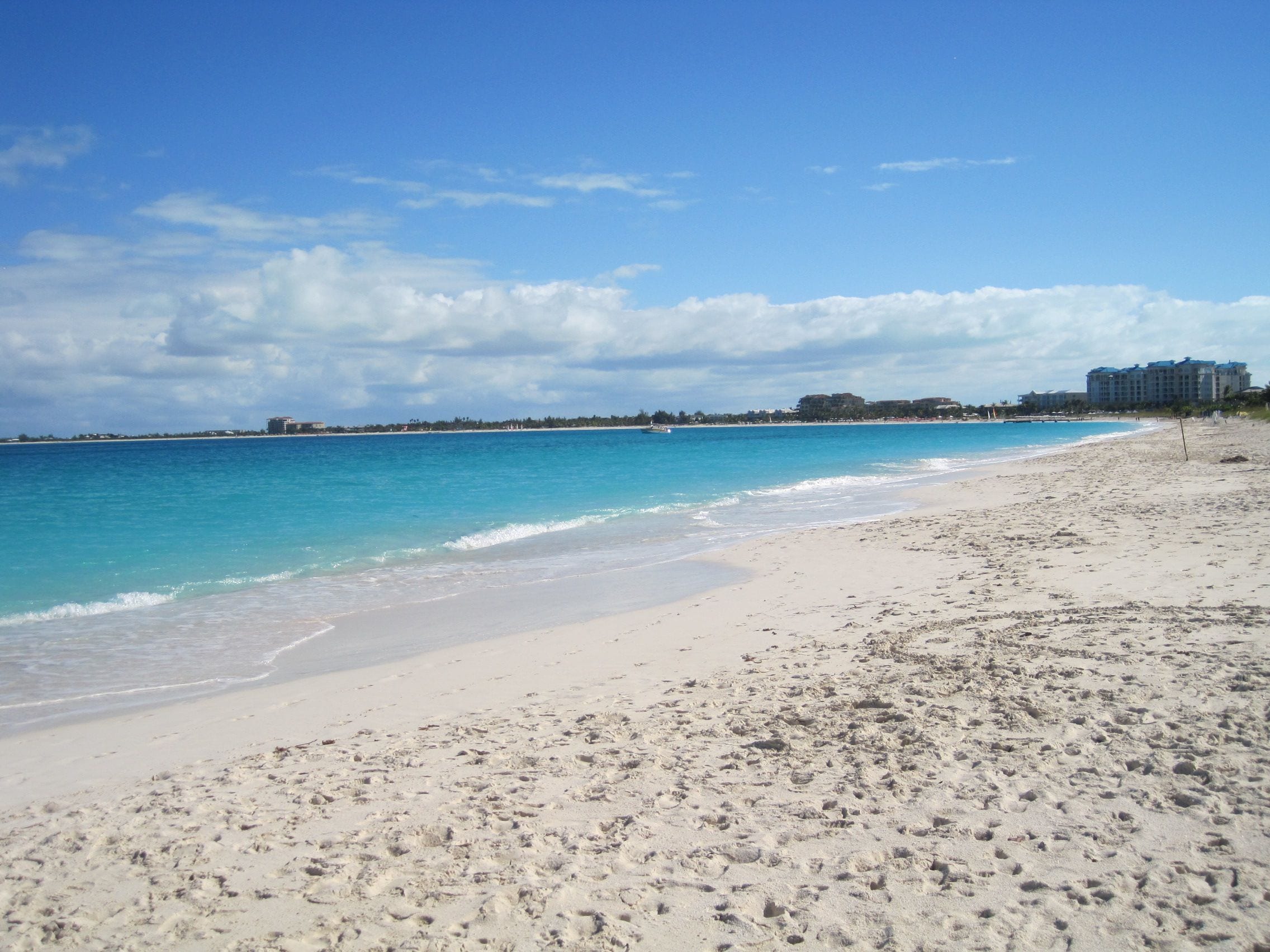 White sand curving along the shore at Grace Bay under a blue sky, the shallow water grading from pale turquoise to deep blue and the low Providenciales resort strip in the distance