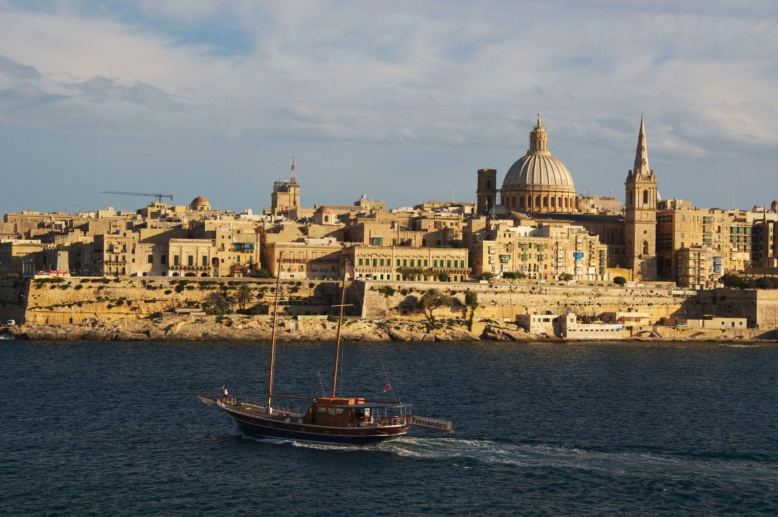 Fort St Angelo and the Three Cities across the turquoise water of the Grand Harbour, seen from the Valletta bastions with a traditional Maltese boat in the foreground