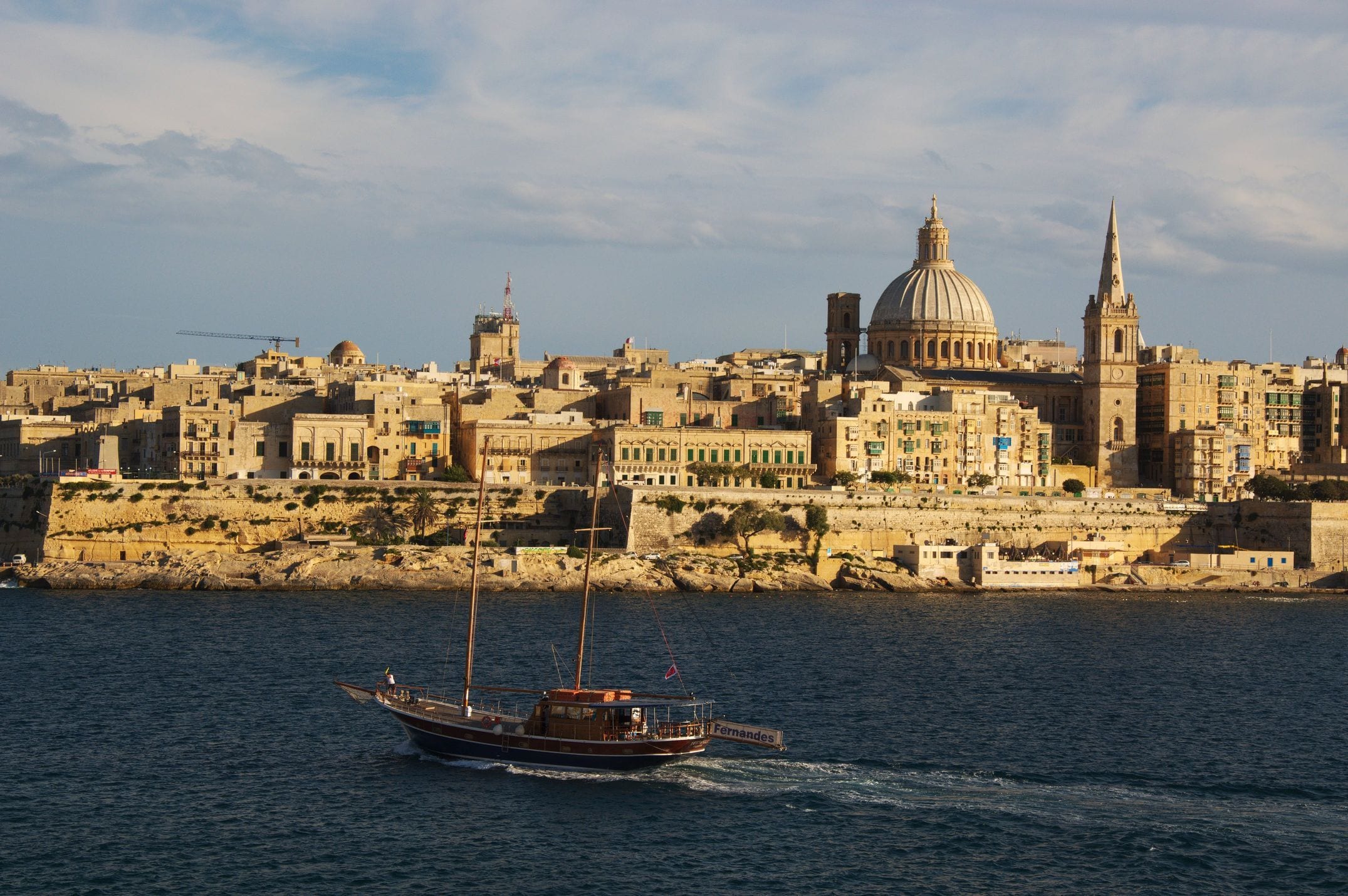 Fort St Angelo and the Three Cities across the turquoise water of the Grand Harbour, seen from the Valletta bastions with a traditional Maltese boat in the foreground
