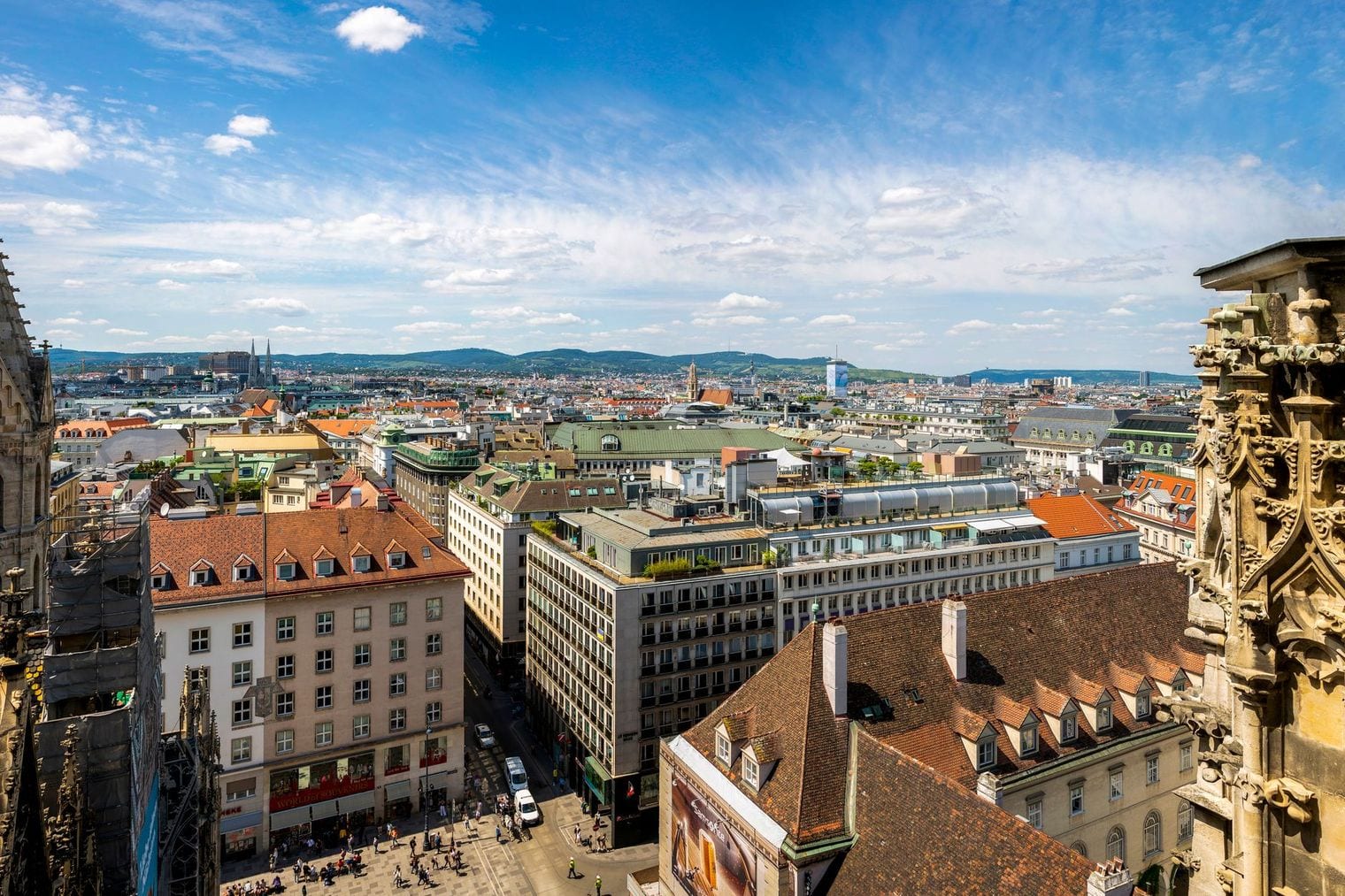 View over Vienna's red-tiled old town rooftops from the tower of St. Stephen's Cathedral, with carved Gothic stonework in the foreground and the Vienna Woods on the horizon