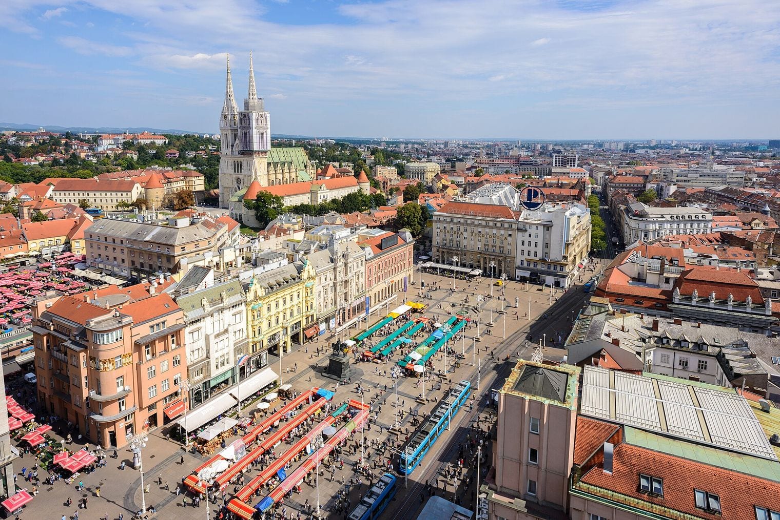 Aerial view over Ban Jelacic Square in Zagreb, with a blue tram crossing the plaza, the red parasols of Dolac market to the left and the twin spires of Zagreb Cathedral rising behind the red-tiled rooftops.